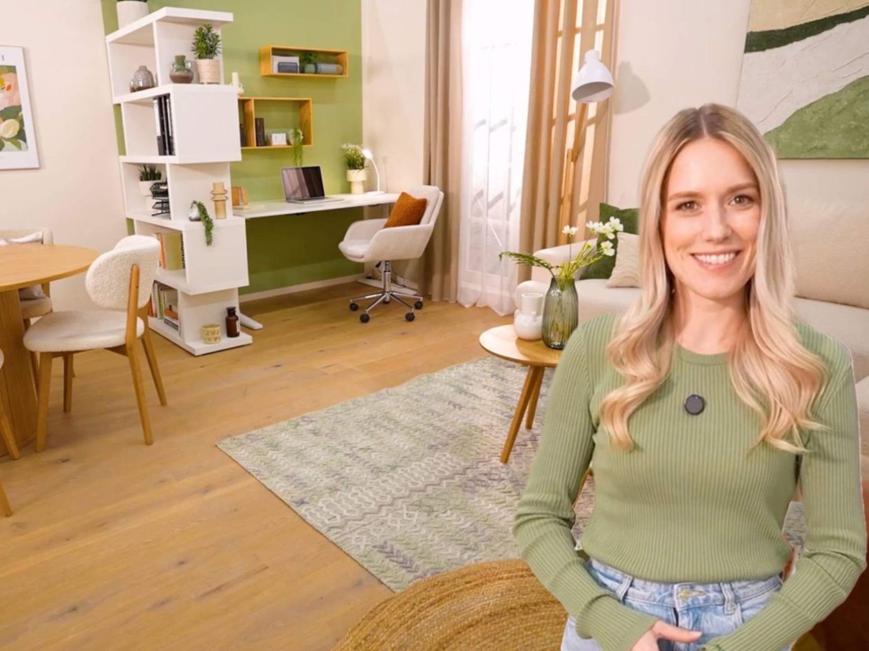 Lächelnde Frau in einem gemütlichen Wohnzimmer mit grünen Akzenten, einem Schreibtisch, Stühlen und Regalen. Der helle Holzboden und das natürliche Licht werten den Raum auf.