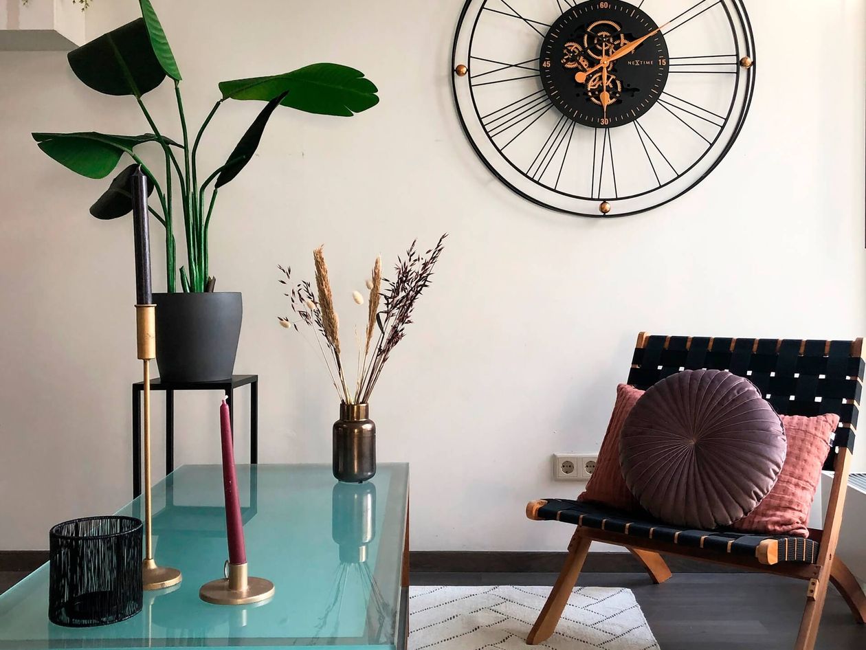 Modernes Wohnzimmer mit einem Glastisch, Kerzen, Topfpflanzen, einer großen Wanduhr und einem Stuhl mit rosa und lila Kissen.