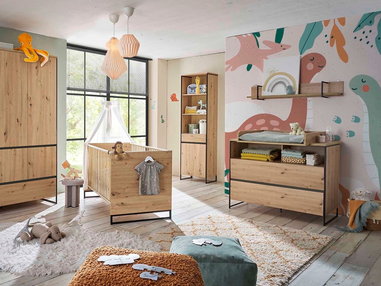Vollständig eingerichtetes Babyzimmer mit naturholzfarbenem Möbelset (Kleiderschrank, Babybett mit Himmel, Wickelkommode und Hochregal) vor bunter Dino-Tapete, zwei Pendelleuchten an der Decke.
