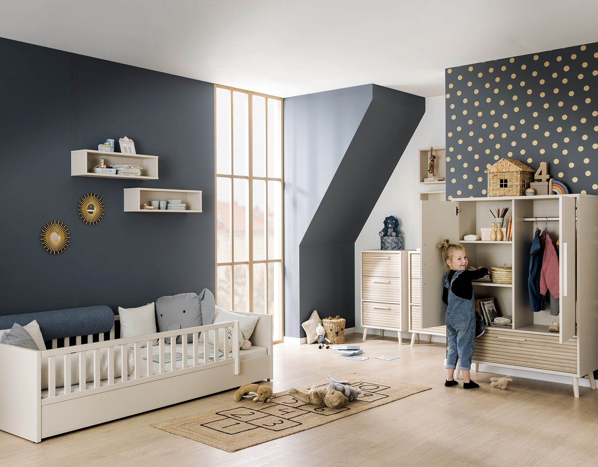 Ein kleines Mädchen steht an einem offenn Schrank in einem Kinderzimmer blauen Wänden und gelben Punkten und verspielter Deko. Eingerichtet ist es mit den beigen Möbeln der Paidi Serie Eefje mit Absetzungen in Eiche in Lamellenoptik und dem bodennahen Bett Little Flo in Weiß..