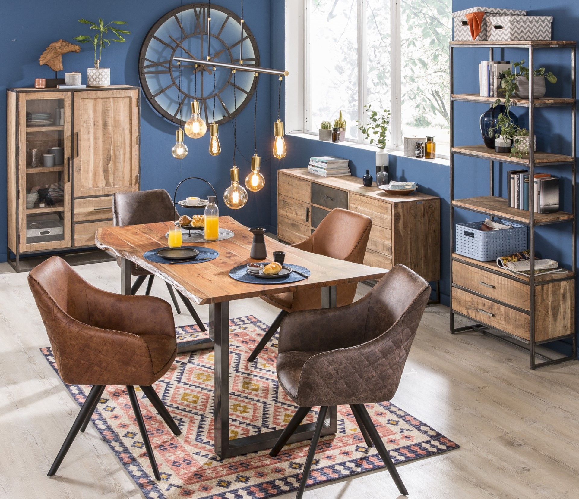 Esszimmer mit einem Holztisch, vier Stühlen, einer großen Uhr, Pendelleuchten sowie mehreren Regalen und Kommoden.