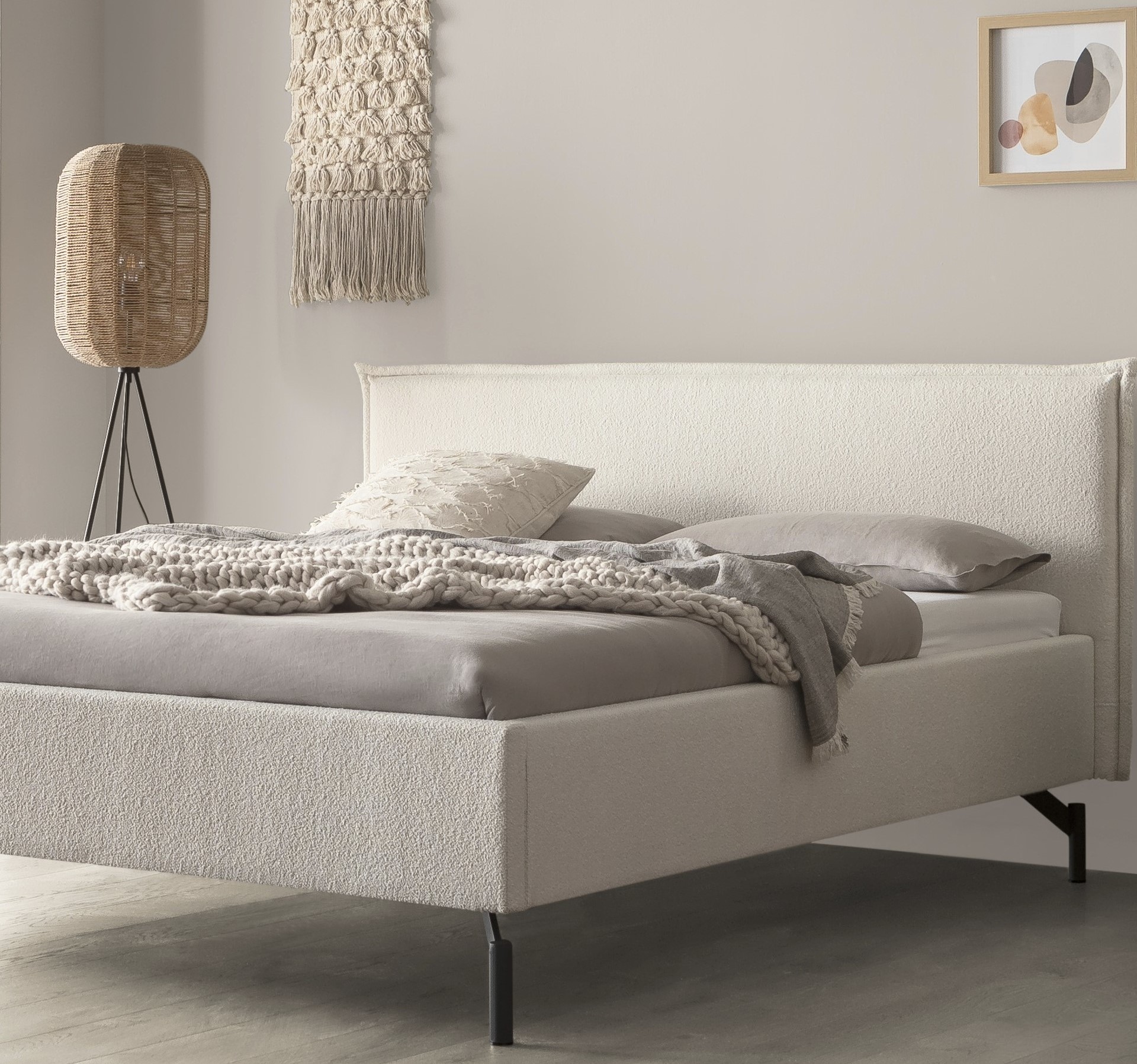 Minimalistisches Schlafzimmer mit einem beigen Polsterbett, grauer Bettwäsche, geflochtener Lampe und einer hellgrauen Wand.