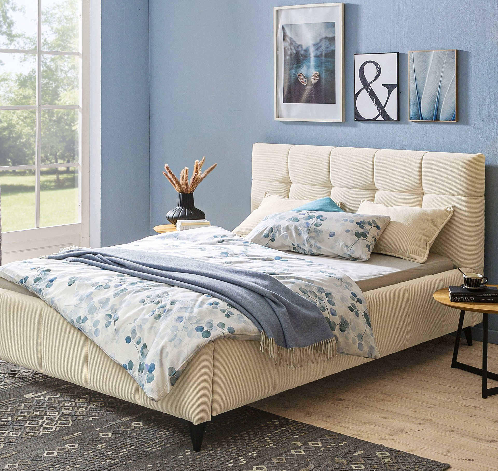 Gemütliches Schlafzimmer mit einem cremefarbenen Polsterbett, blauer und weißer Bettwäsche, dekorativen Kissen und einem Beistelltisch.