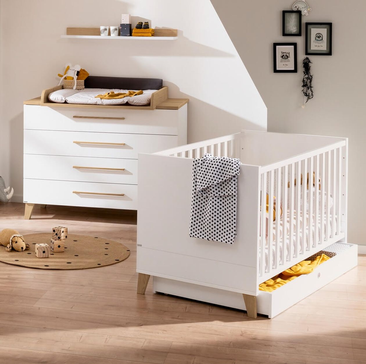 Helles Babyzimmer mit Möbeln in Weiß und Holzoptik. Im Vordergrund steht ein Gitterbett, im Hintergrund eine Wickelkommode und ein großer Schrank. Der Boden wirkt freundlich und es sind Kuscheltiere sowie pastellfarbene Deko zu sehen.