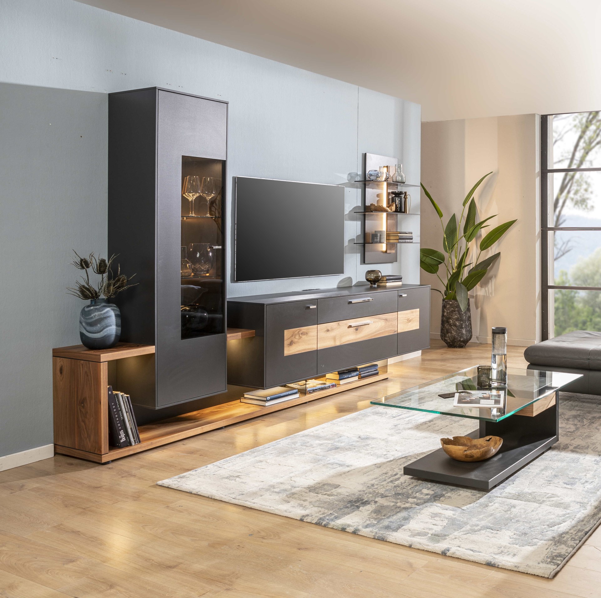 Modernes Wohnzimmer mit eleganter Wohnwand. Dazu TV, Couchtisch aus Glas und dekorativen Pflanzen. Große Fenster lassen natürliches Licht in den Raum.
