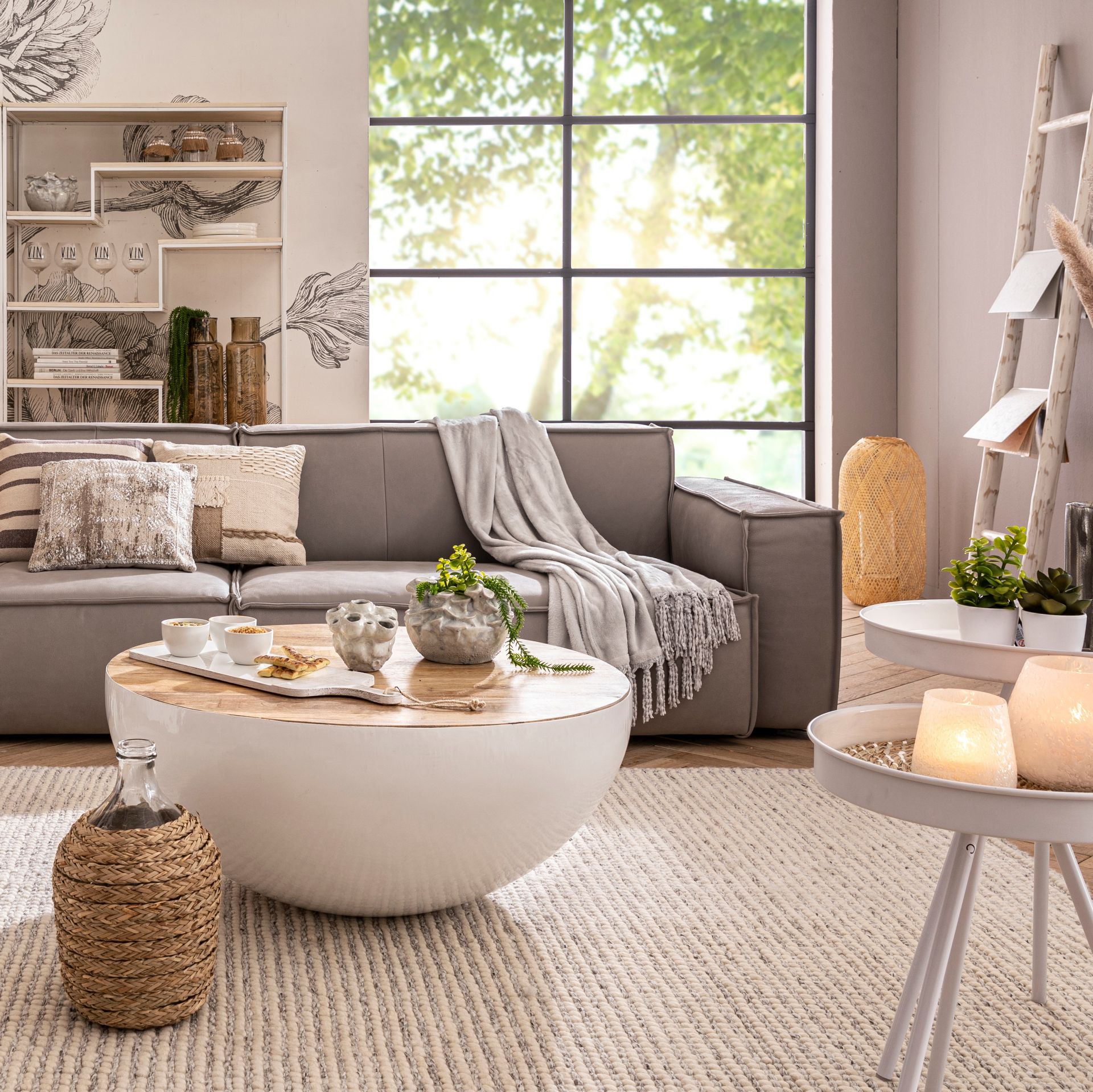 Im Hintergrund befinden sich ein weißes Metallregal, ein großes Fenster und ein graues Sofa. Davor liegt ein cremefarben-grauer Teppich mit Rillenoptik, auf dem drei weiße Couchtische stehen. Der Hintere sieht aus wie die Hälfte einer weißen Kugel mit Holzplatte und die Vorderen sind ein Set bestehend aus zwei weißen Metall-Beistelltischen mit jeweils drei dünnen Beinen.