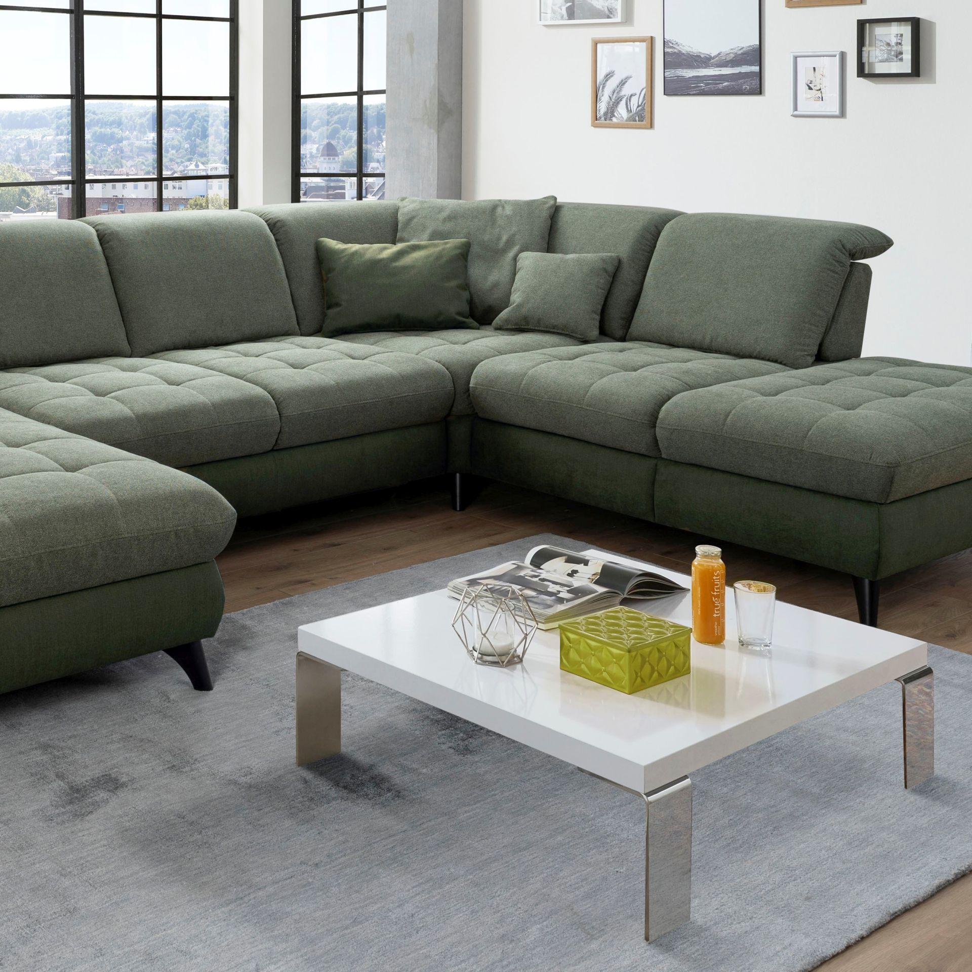 Auf dem Foto befindet sich ein dunkelgrünes U-förmiges Sofa auf einem hellgrauen Teppich mit einem davorstehenden quadratischen Couchtisch mit silber glänzenden Beinen und einer weißen Hochglanz-Platte.