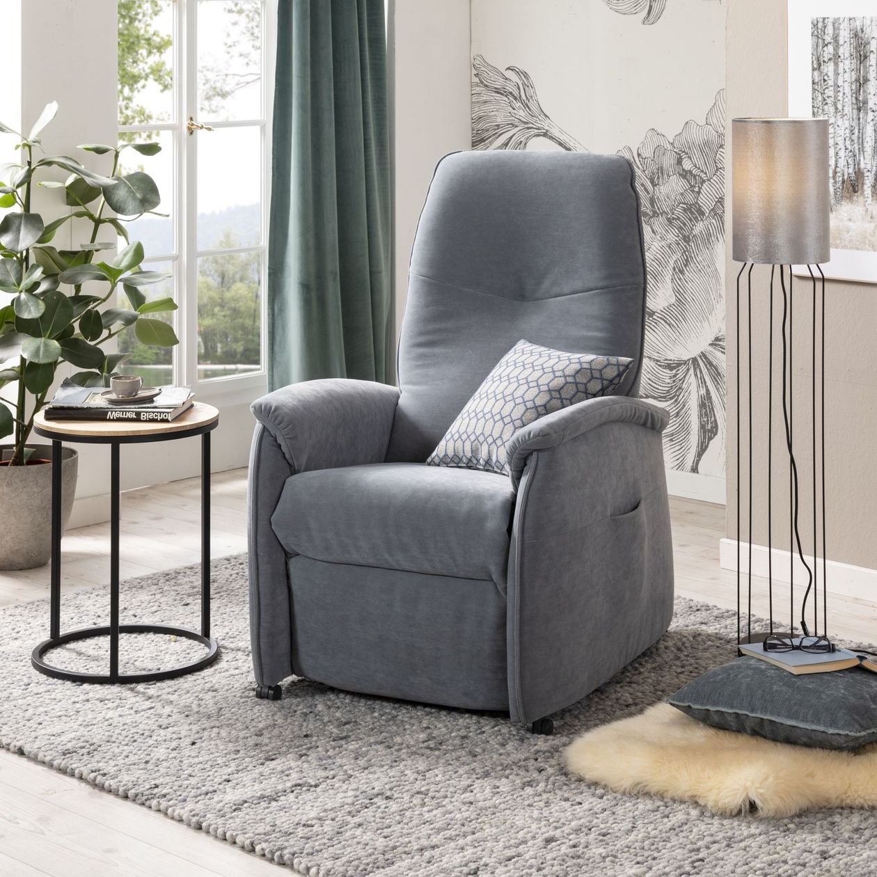 Auf dem Foto befindet sich ein blau-grauer Relaxsessel mit Mikrofaser-Bezug und Aufstehfunktion. Er steht auf einem grauen Teppich, rechts daneben eine Stehlampe und links daneben ein Beistelltisch mit schwarzen Metallbeinen.