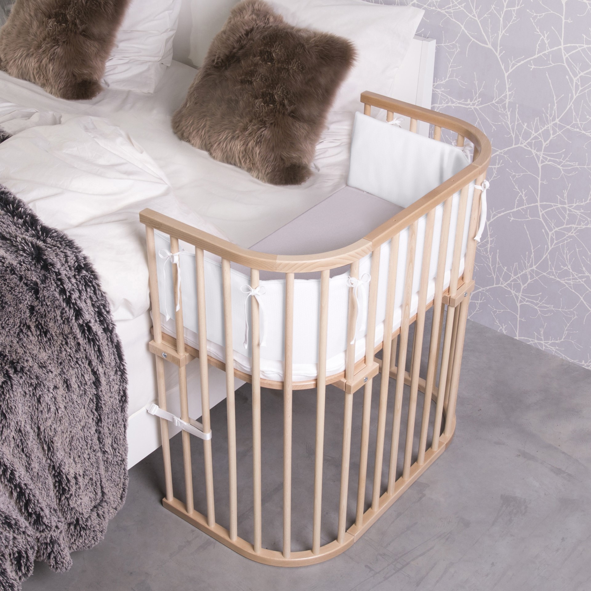 Beistellbett Babybay Boxspring der Marke Babybay aus naturbelassenem Holz neben einem kuschelig bezogenen Bett mit Fellkissen und Felldecke.