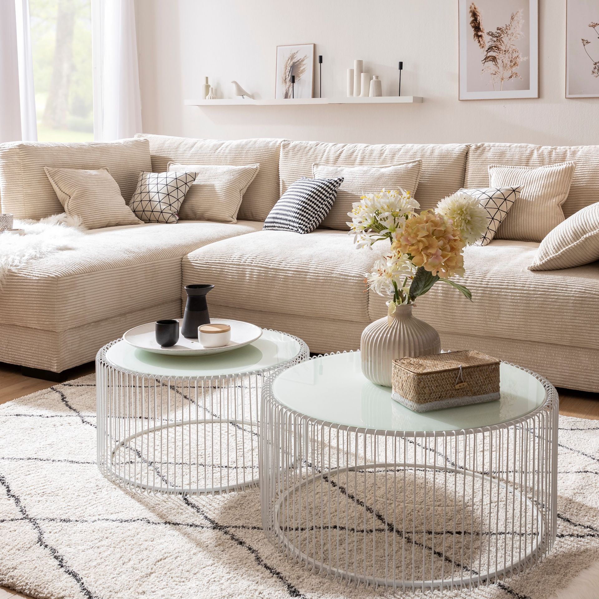 Im Hintergrund befindet sich ein cremefarbenes Cord-Sofa mit hellen Kissen auf einem hellen Teppich mit dunklem Rautenmuster. Vor dem Sofa steht ein Satz Couchtische bestehend aus zwei runden Tischen in Weiß, welche ein Gestell aus dünnen Metallstäben besitzen und eine milchige Glasplatte.