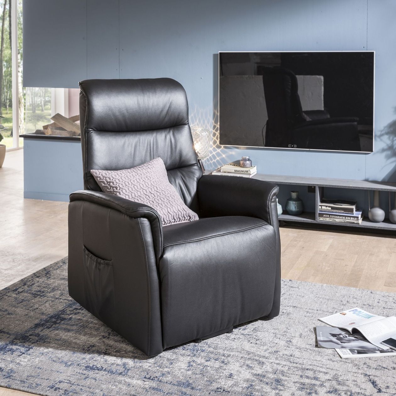 Auf dem Bild befindet sich ein schwarzer Leder-Relaxsessel mit Aufstehhilfe, welcher auf einem grau-blauen Teppich steht. Die Wand mit einem Fernseher im Hintergrund ist blau gestrichen.