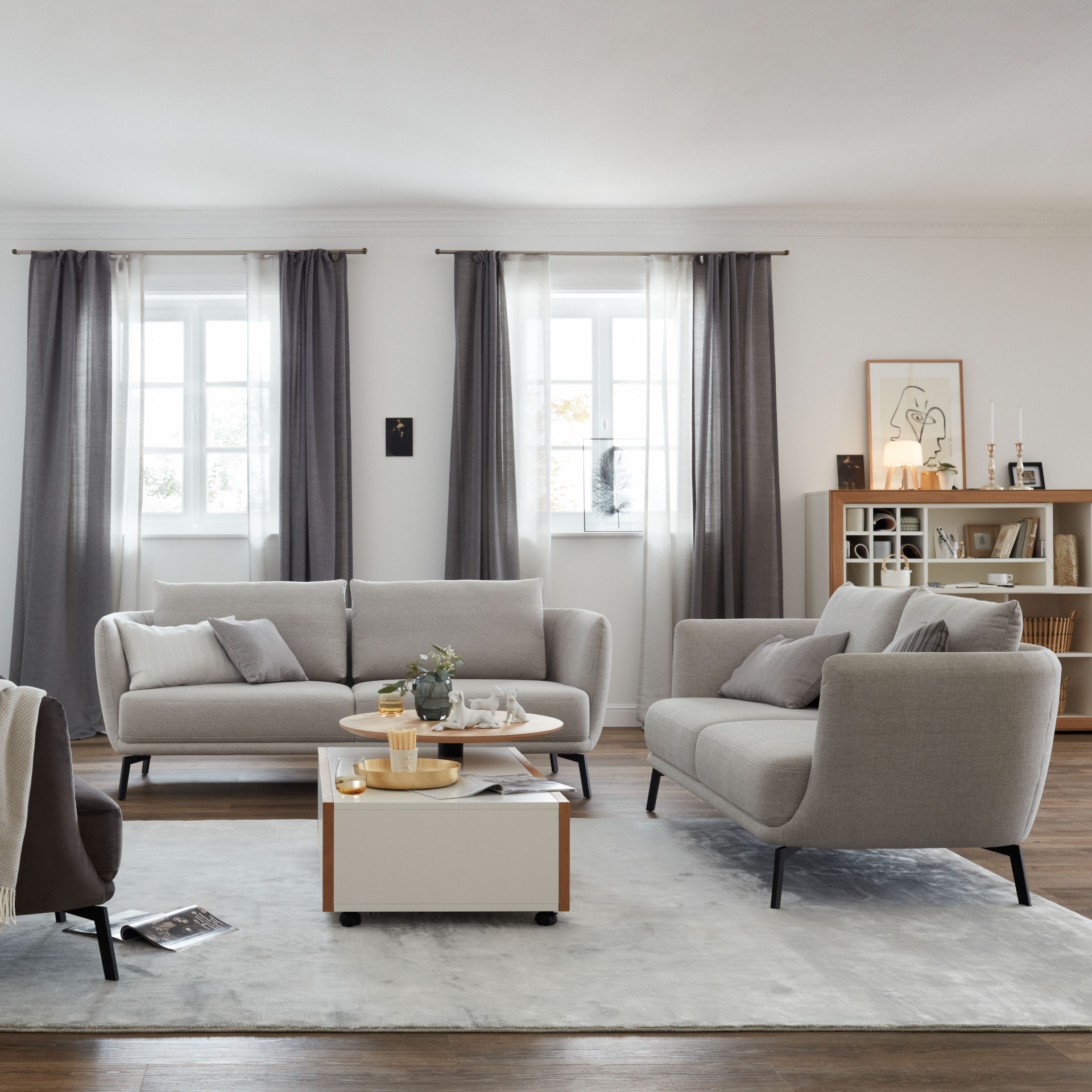 Um einen weißen Couchtisch mit rundem Holzaufsatz stehen zwei graue Sofas des Typs Pearl der Marke SCHÖNER WOHNEN-Kollektion mit schwarzen Füßen.