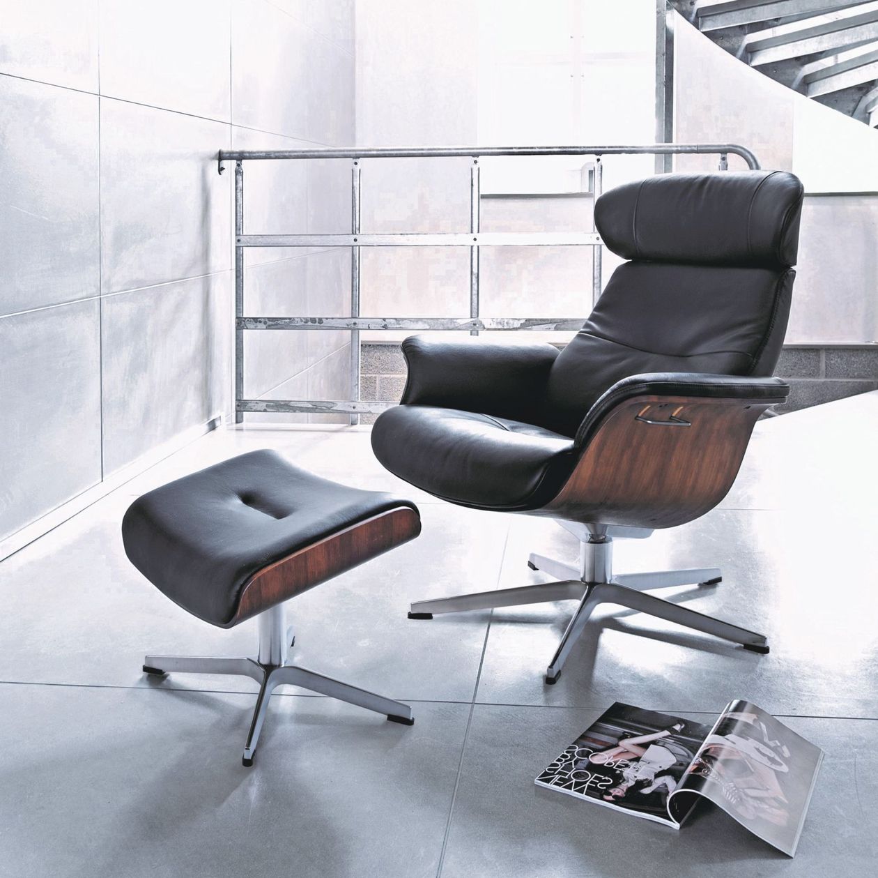 Ein moderner schwarzer Relaxsessel aus Leder mit Holzelementen und einem verchromten Standfuß, daneben ein passender Fußhocker. Der Raum ist hell und modern gestaltet mit grauen glänzenden Fliesen und einem Treppengeländer aus Metall im Hintergrund.