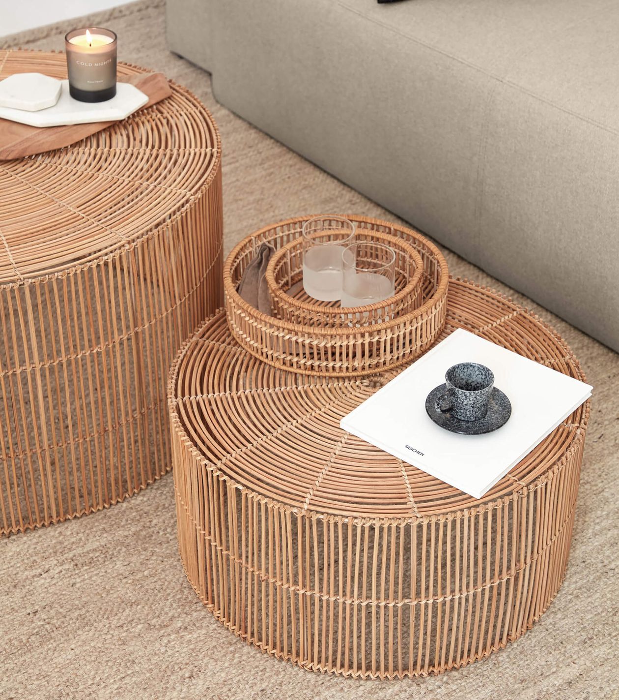 Zwei Korbtische mit Dekoartikeln: eine brennende Kerze, Untersetzer, ein Buch und eine Tasse. Im Hintergrund ist teilweise ein beiges Sofa zu sehen.