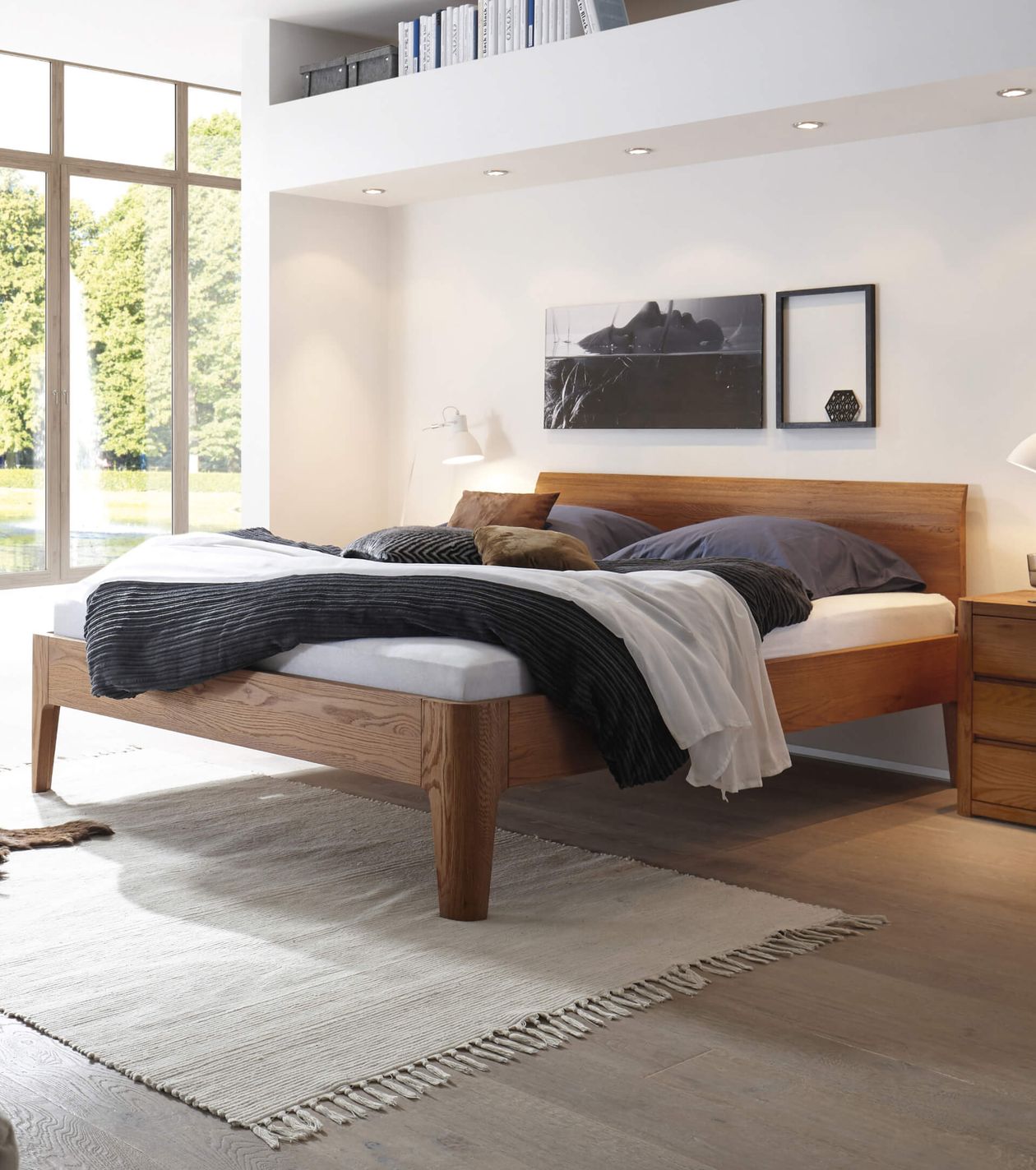 Modernes Schlafzimmer mit Holzbett, weißen Wänden und großen Fenstern. Bett ist mit dunkler und heller Bettwäsche bedeckt, beige Teppich am Boden.