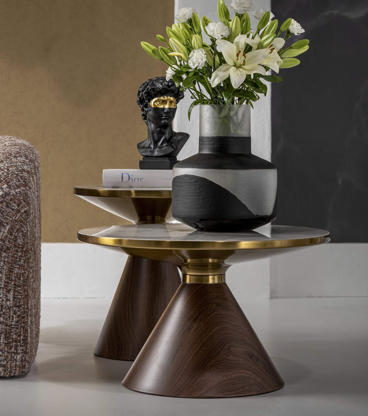 Moderne runde Tische mit Akzenten aus Holz und Messing, einer mit einer Vase voller Lilien, der andere mit einer Büste und Büchern, vor einem minimalistischen Hintergrund.