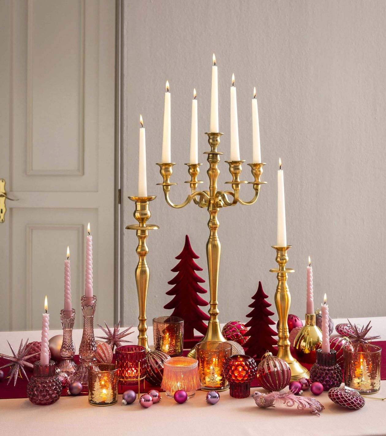 Eine elegante Tischdekoration mit hohen goldenen Kerzenleuchtern, brennenden Kerzen sowie roten und rosa Weihnachtsdekorationen, darunter kleine Tannenbäumchen und Christbaumkugeln.