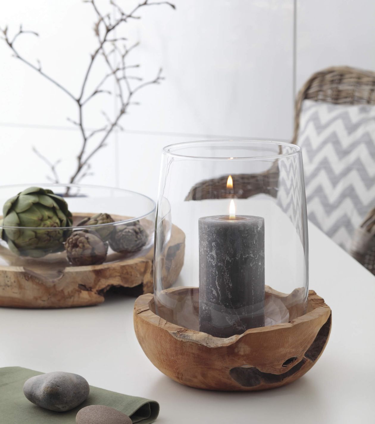 Eine brennende graue Kerze in einem Glashalter mit Holzsockel, neben einer Schale mit getrockneten Artischocken, auf einem Tisch mit grünem Tischtuch und Steinen.