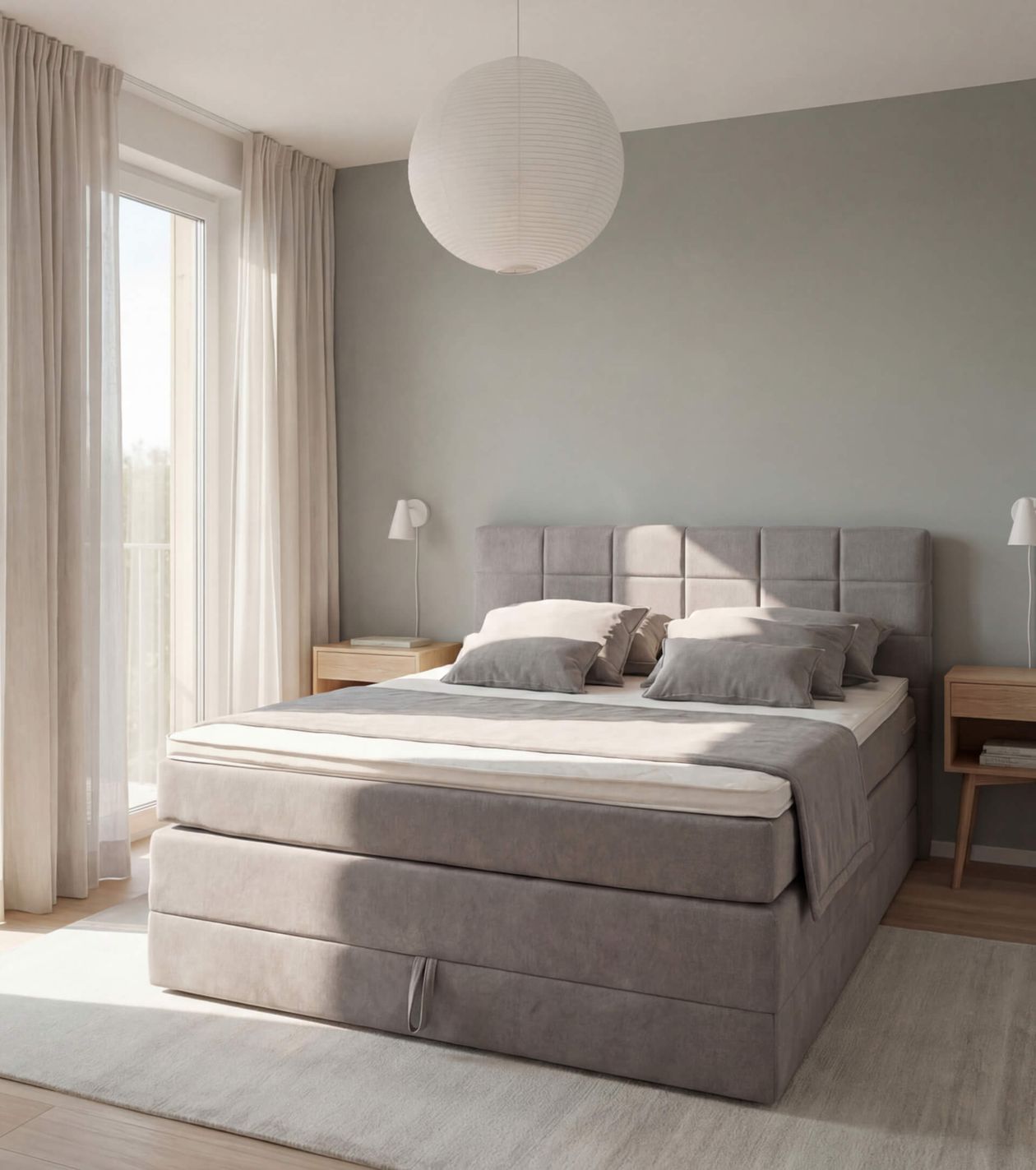 Modernes Schlafzimmer mit grauem Boxspringbett, zwei Nachttischen, hellen Vorhängen und einer großen, runden Hängelampe.