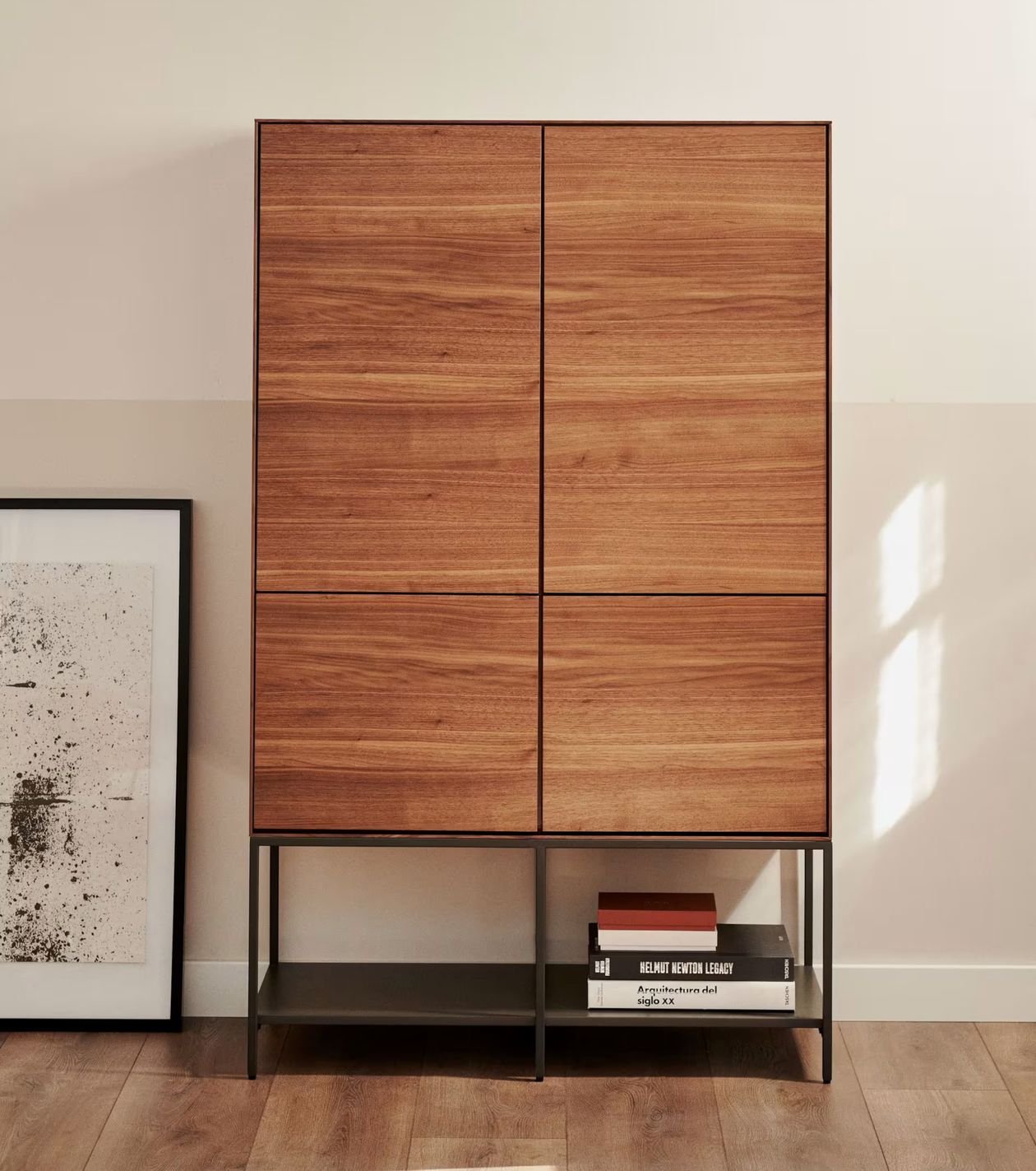Holzschrank mit vier quadratischen Paneelen auf Metallbeinen, einem schwarz-weiß gerahmten Kunstwerk, das an seiner Seite lehnt, und zwei Büchern darunter.