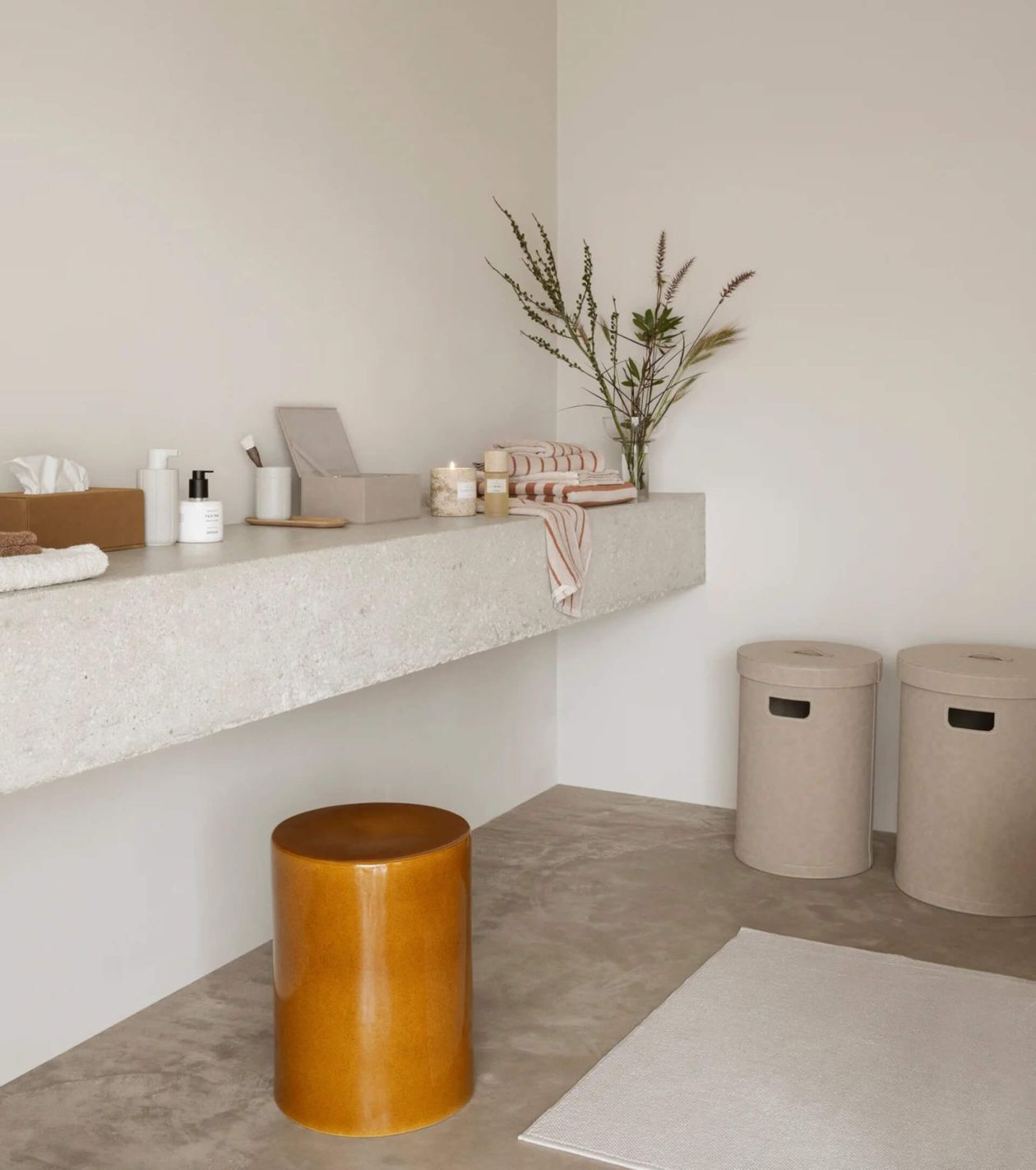 Minimalistisches Bad mit Betonwaschbecken, braunem Hocker, zwei runden Wäschekörben und dekorativen Pflanzen auf der Ablage.