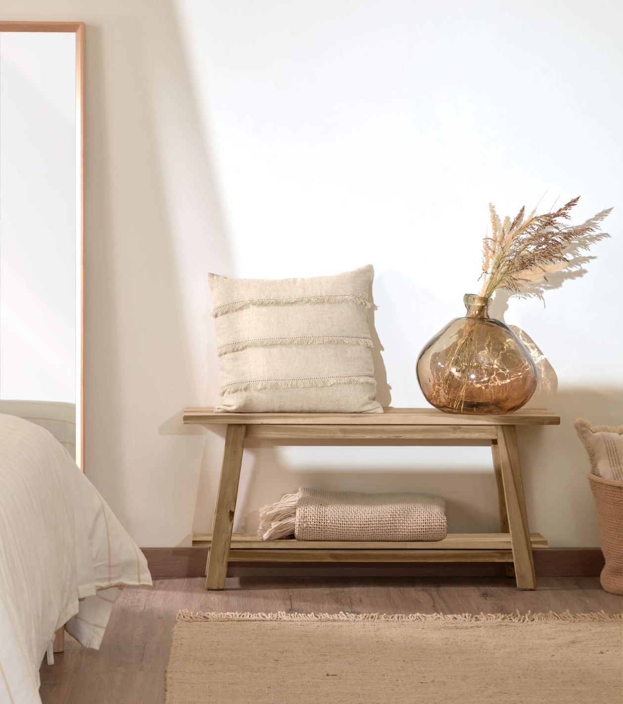 Holzbank mit einem beigen Kissen und einer Glasvase mit getrockneten Pflanzen. Darunter befinden sich ein geflochtener Korb und eine gefaltete Decke in einem minimalistisch eingerichteten Raum.