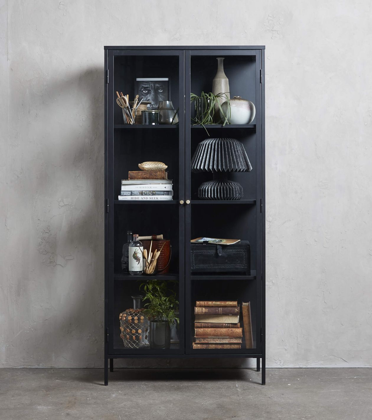 Schwarze Metallvitrine mit Glastüren, in der Bücher, Dekoartikel, eine Pflanze, Körbe und eine kleine Büste auf einem Betonhintergrund zu sehen sind.