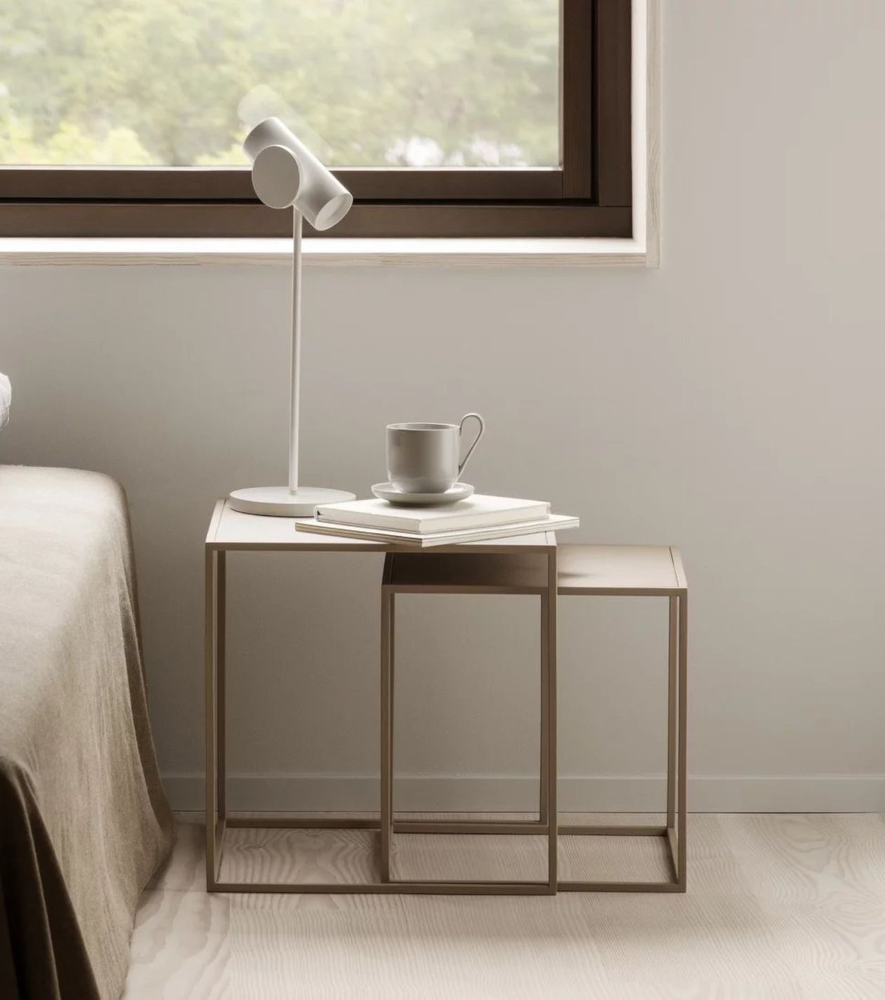 Minimalistische Einrichtung am Bett mit gestapelten Tischen, einer weißen Tasse auf einer Untertasse, einem geschlossenen Buch und einer modernen Lampe in der Nähe eines Fensters.