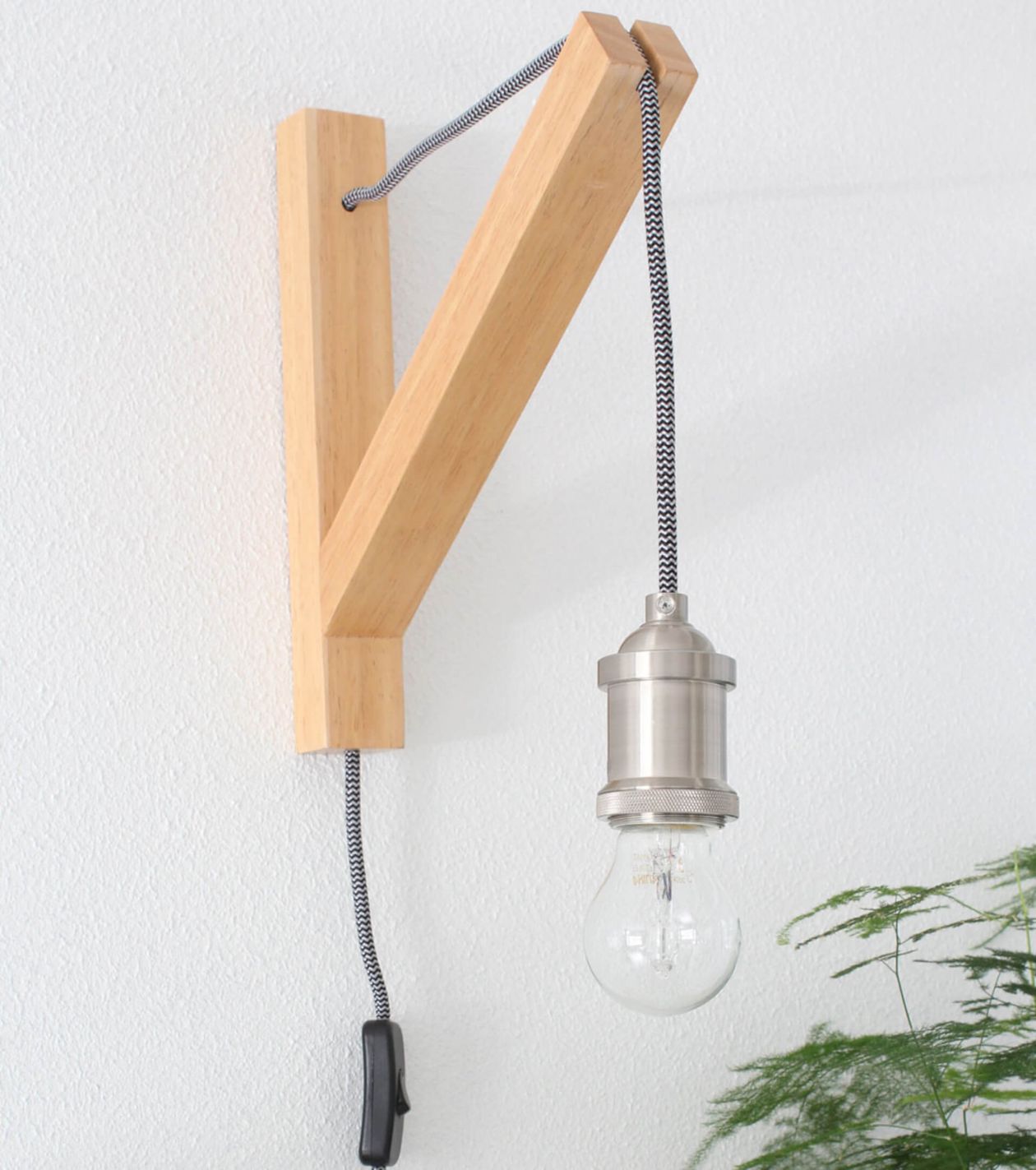 Wandleuchte aus Holz vor einer weißen Wand mit einer sichtbaren Glühbirne und einer grünen Farnpflanze daneben.