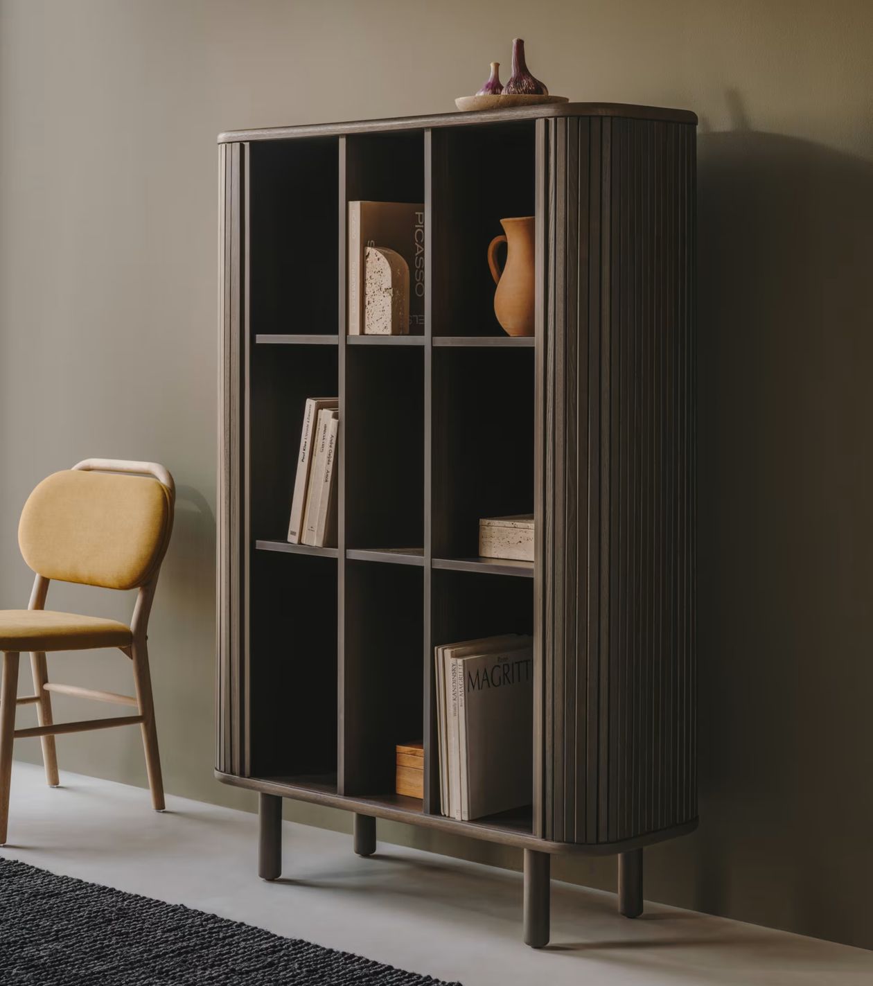 Modernes Bücherregal aus Holz mit neun Fächern, Dekoartikeln und Büchern. Daneben steht ein senfgelber Stuhl vor einer beigen Wand.
