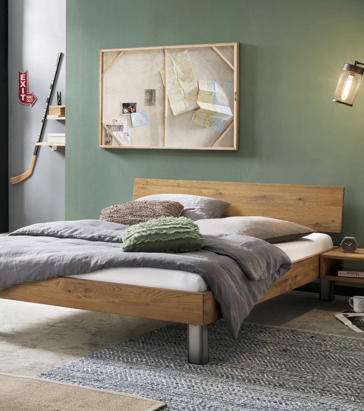 Ein modernes Schlafzimmer mit einem Holzbett, grauer Bettwäsche und grünen Zierkissen. An der grünen Wand hängen eine Pinnwand und ein Hockeyschläger.