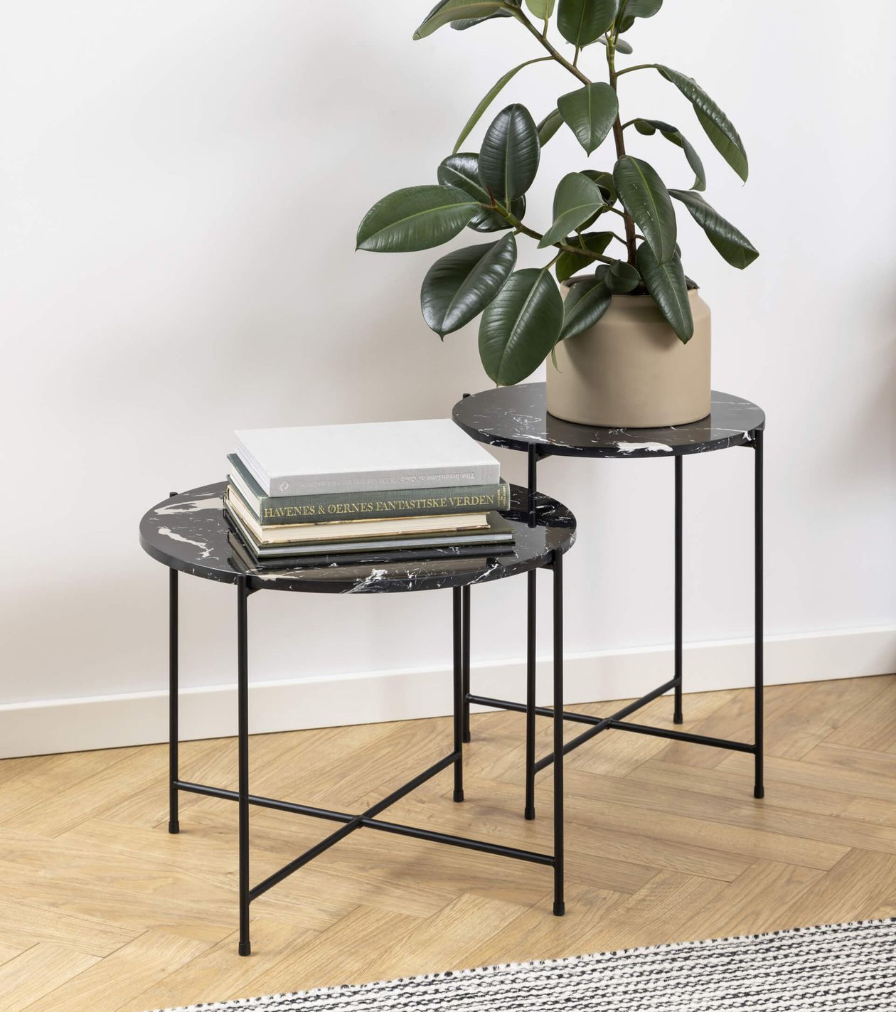 Zwei schwarze Beistelltische mit Marmorplatte und dünnen Metallbeinen; auf dem einen liegt ein Stapel Bücher, auf dem anderen steht eine grüne Topfpflanze, auf hellem Holzboden.