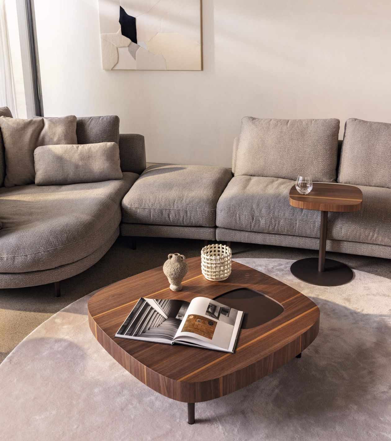 Modernes Wohnzimmer mit grauem Ecksofa, Couchtisch aus Holz mit Büchern, Dekoartikeln und Beistelltisch mit Glas. Neutrale Farbtöne.