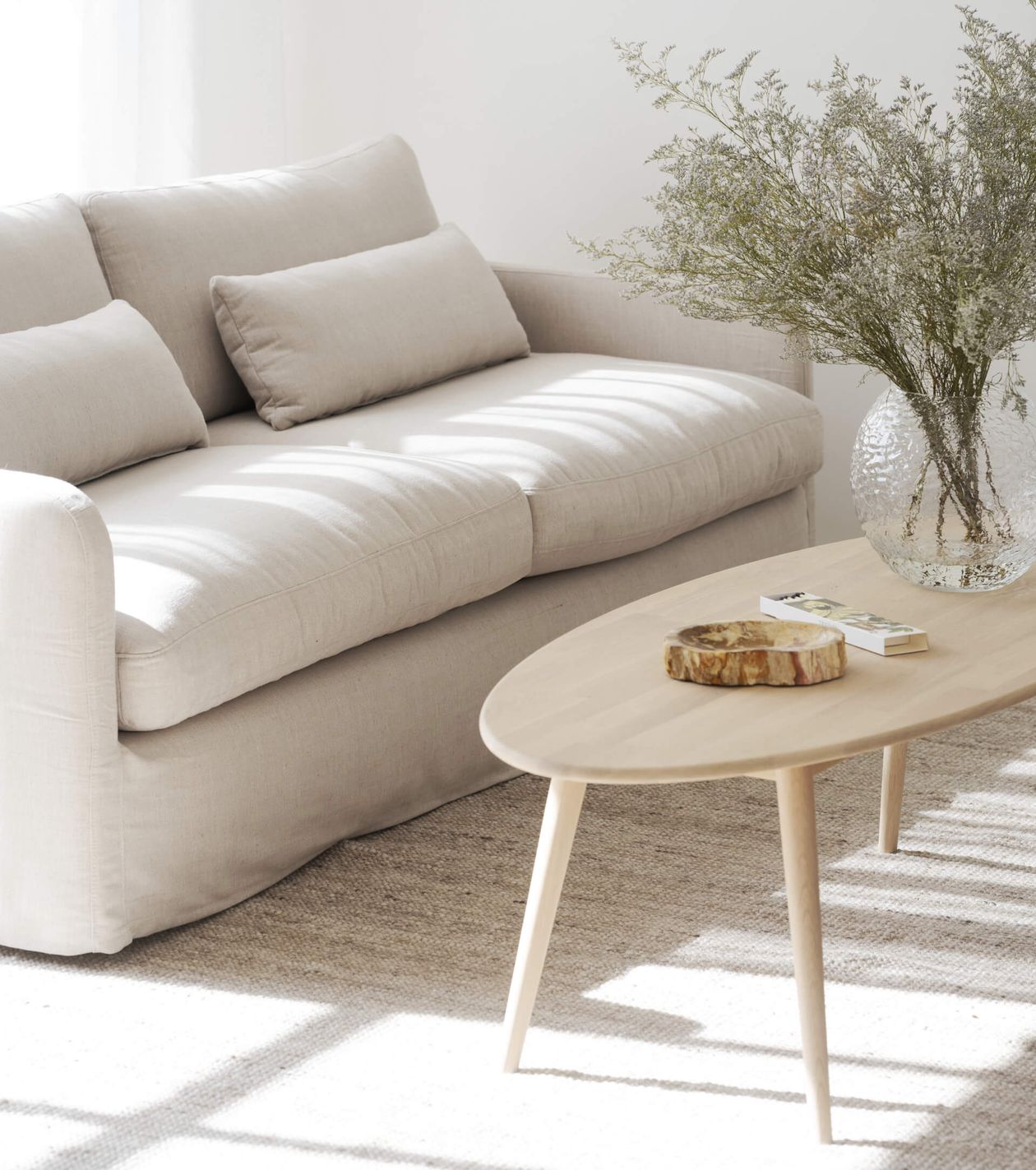 Minimalistisches Wohnzimmer mit einem beigen Sofa, einem Couchtisch aus Holz und einer Glasvase mit Grünpflanzen. Das Sonnenlicht wirft sanfte Schatten auf den Boden.