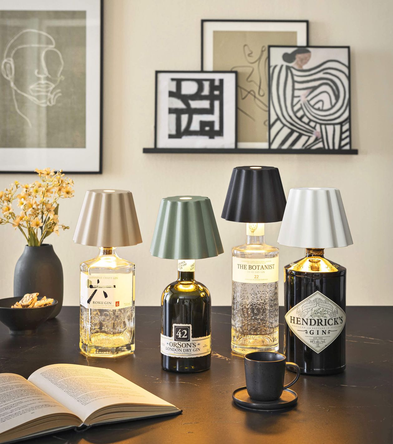 Tischlampen aus Gin-Flaschen mit verschiedenen Lampenschirmen auf einem schwarzen Tisch, umgeben von Kunstwerken, einem Buch, Blumen und einer Tasse.
