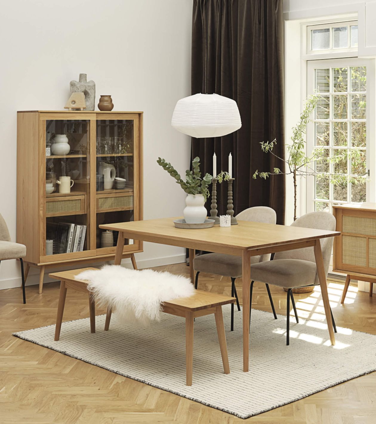 Modernes Esszimmer mit Holztisch, Stühlen und Bank. Vitrine, weiße Pendelleuchte und Pflanzen. Sonnenlicht durch großes Fenster.