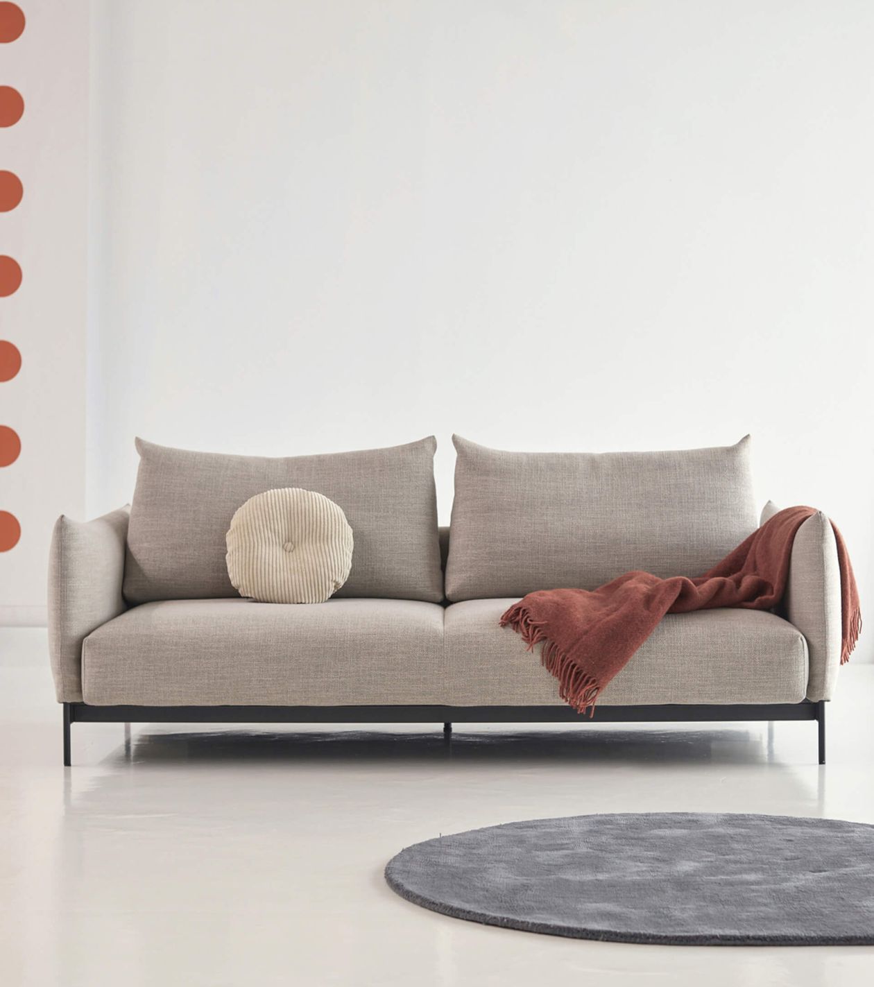 Beiges Sofa mit rundem Kissen und rostfarbener Decke. Grauer Teppich auf hellem Boden, weiße Wand im Hintergrund mit orangen Punkten.