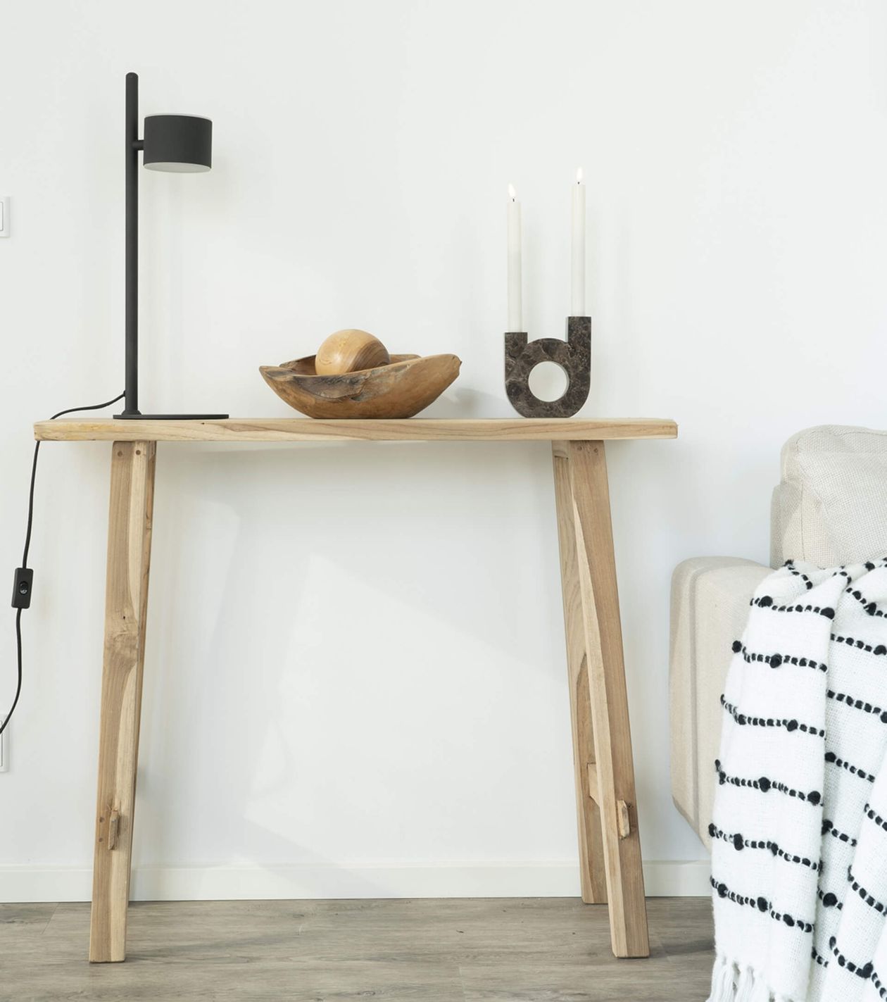 Ein minimalistischer Holztisch mit einer schwarzen Lampe, einer Holzschale und einem modernen Kerzenhalter, daneben ein beiges Sofa mit einer gestreiften Decke.