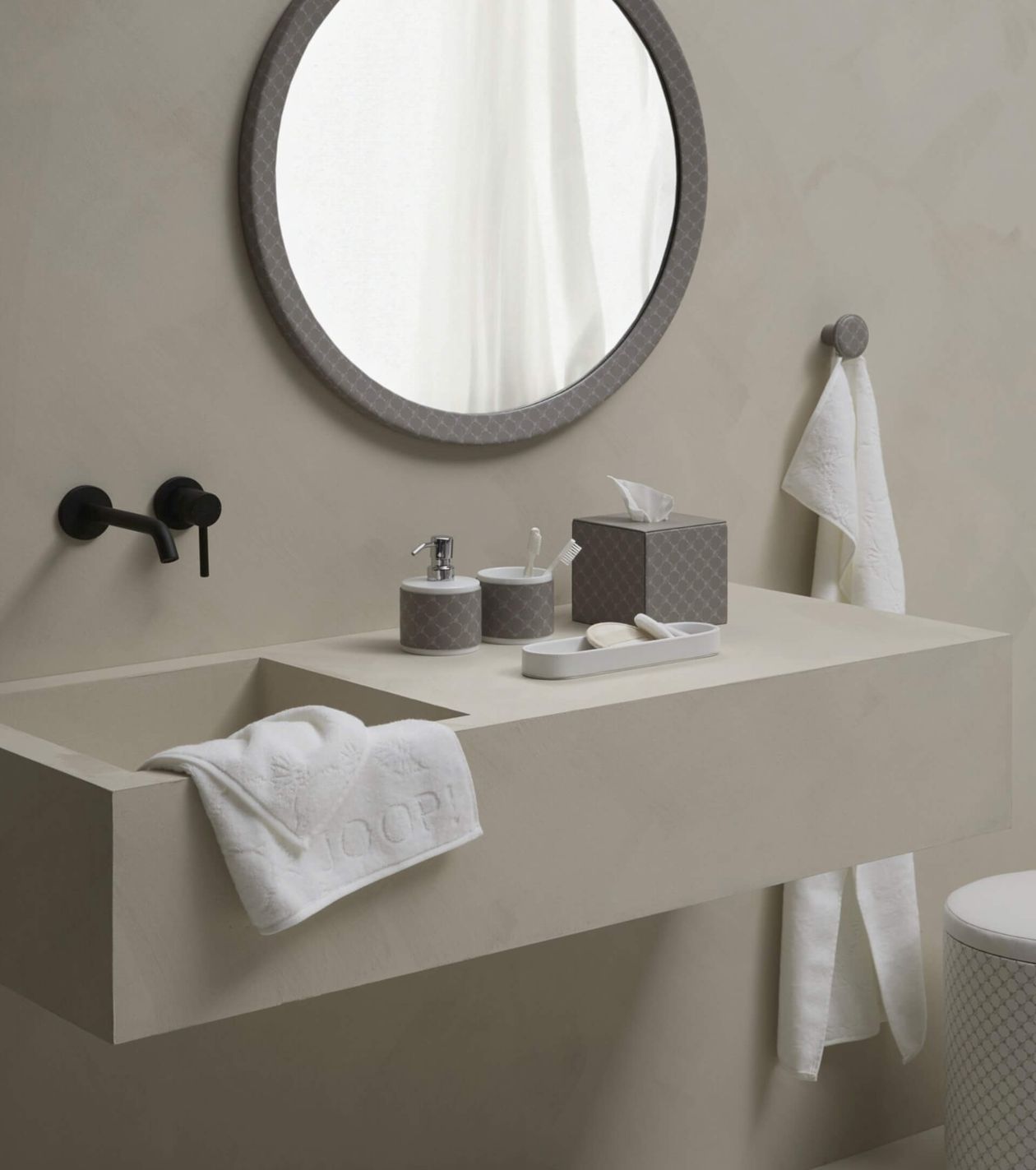 Minimalistisches Badezimmer mit rundem Spiegel, beigem Waschbecken, schwarzem Wasserhahn und ordentlich angeordneten Toilettenartikeln. Handtücher hängen an der Wand und am Waschbecken.