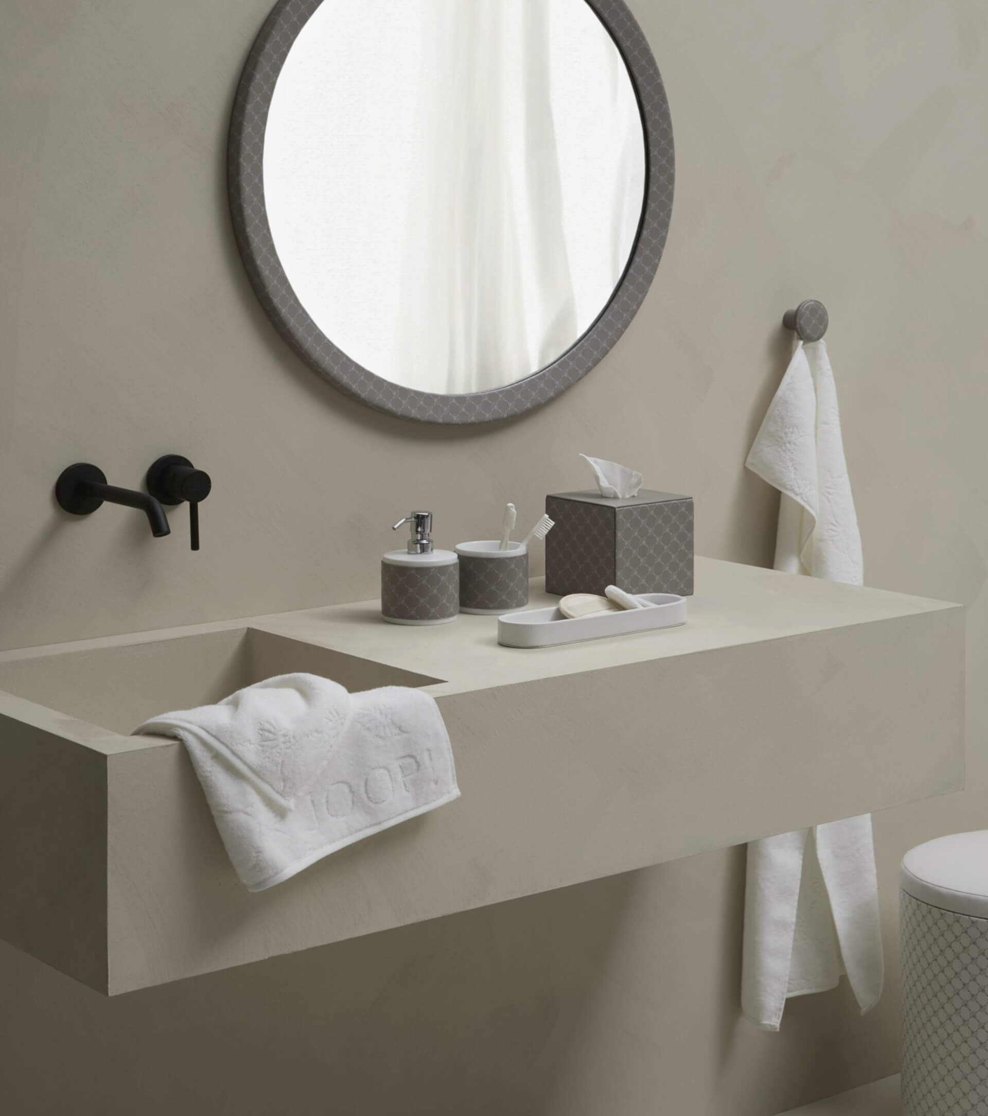 Minimalistisches Badezimmer mit rundem Spiegel, beigem Waschbecken, schwarzem Wasserhahn und ordentlich angeordneten Toilettenartikeln. Handtücher hängen an der Wand und am Waschbecken.