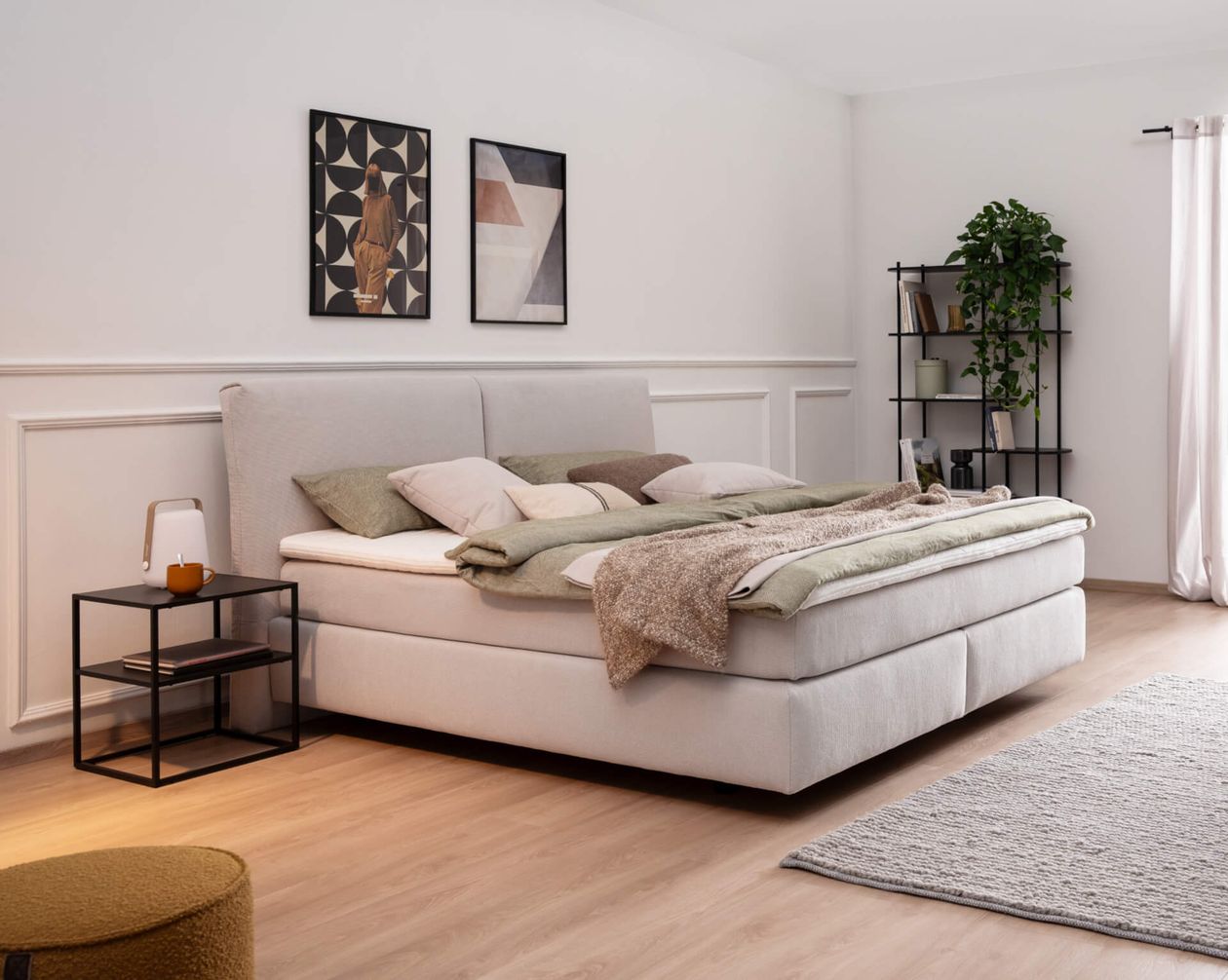 Modernes Schlafzimmer mit einem großen Bett, mehrlagigen Decken, abstrakter Kunst an weißen Wänden, einem kleinen Beistelltisch und einem hohen Regal mit Grünpflanzen.