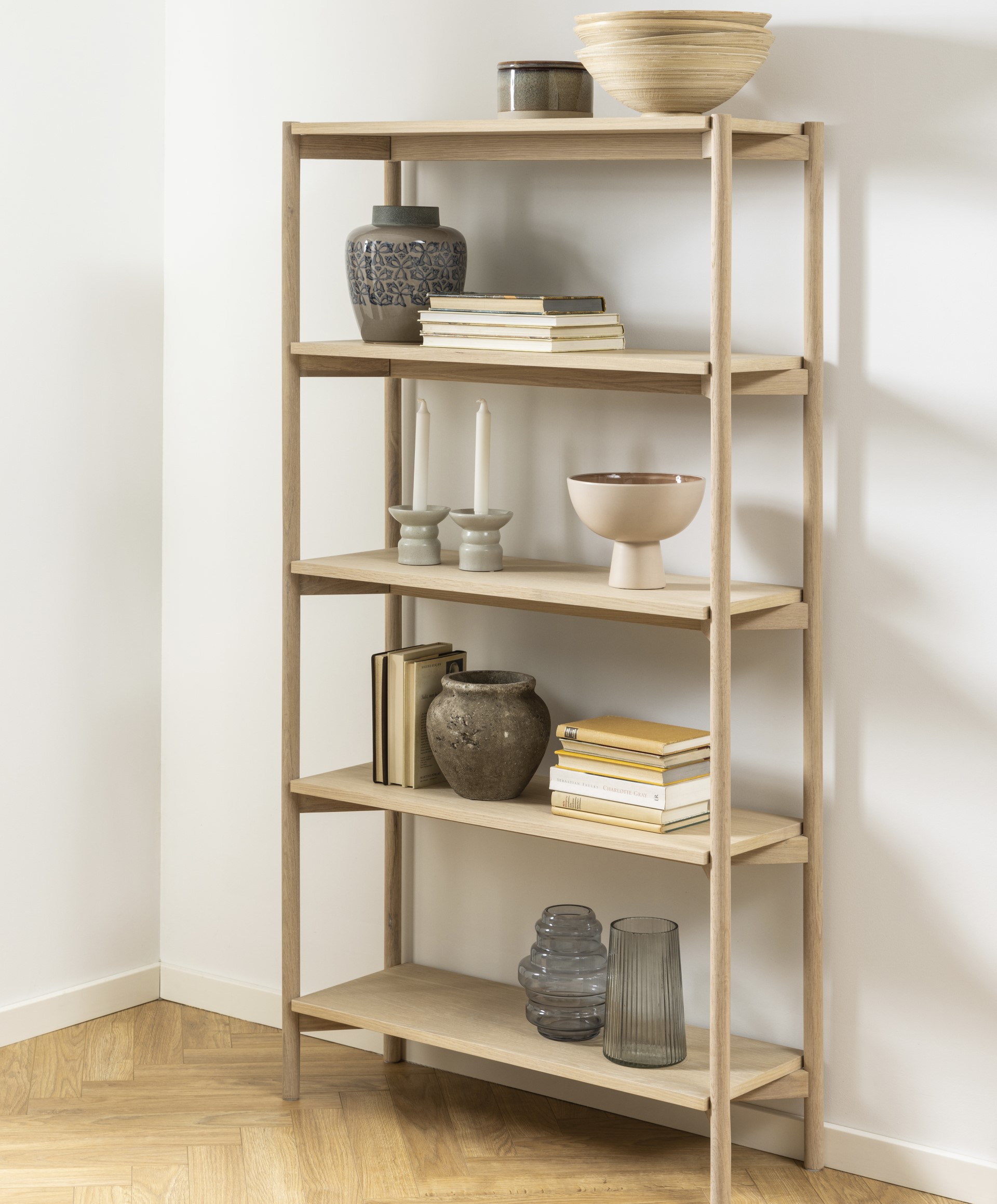 Bücherregal aus Holz mit fünf Regalen, in denen Bücher, Keramikvasen, Kerzen und eine Schale ausgestellt sind, vor einer weißen Wand auf einem Holzboden.