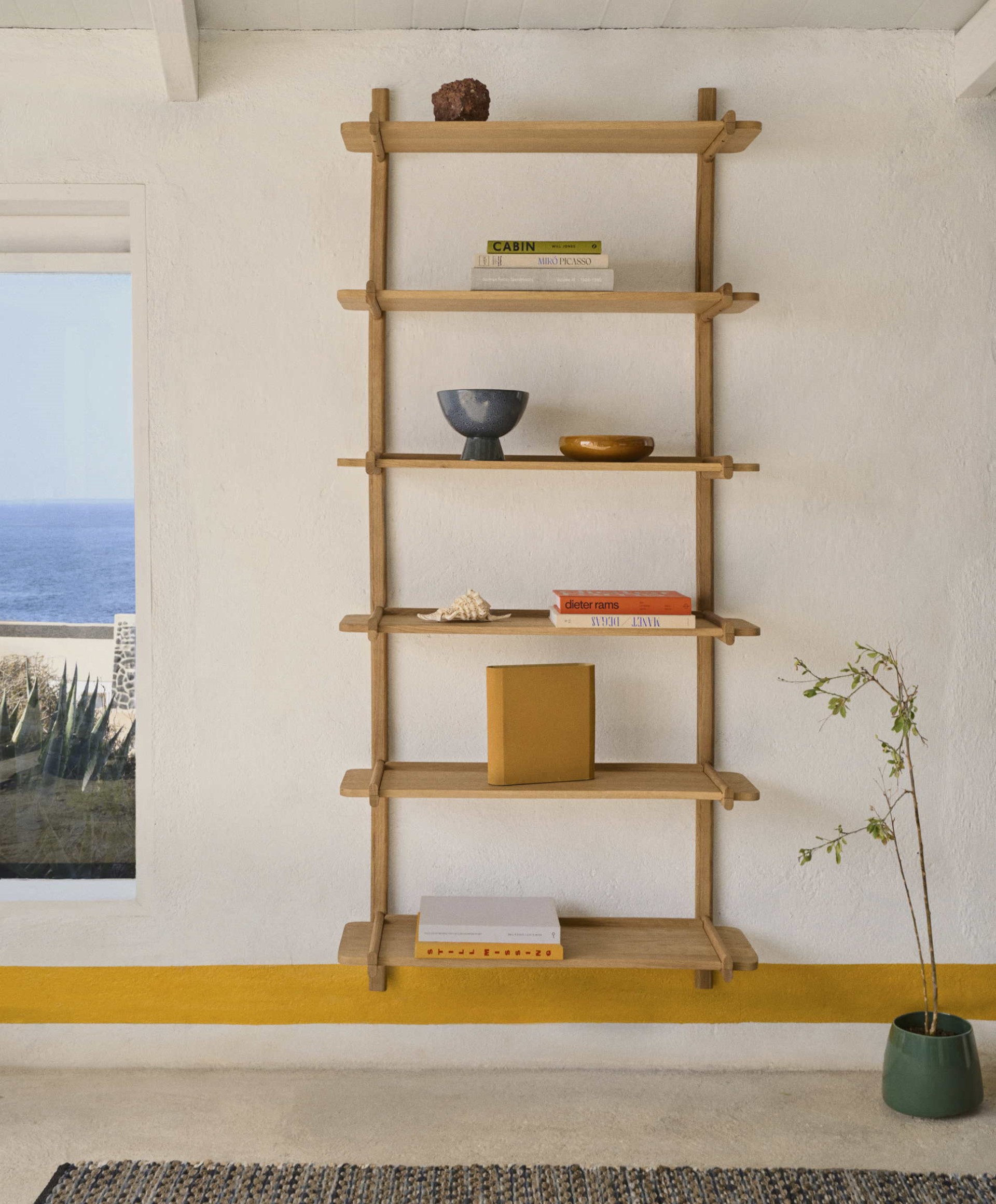 Wandregal aus Holz mit Büchern, einer Schale und Dekorationsgegenständen vor einer weißen Wand. Ein Fenster gibt den Blick auf die Küste frei. Eine kleine Pflanze befindet sich in der Nähe.