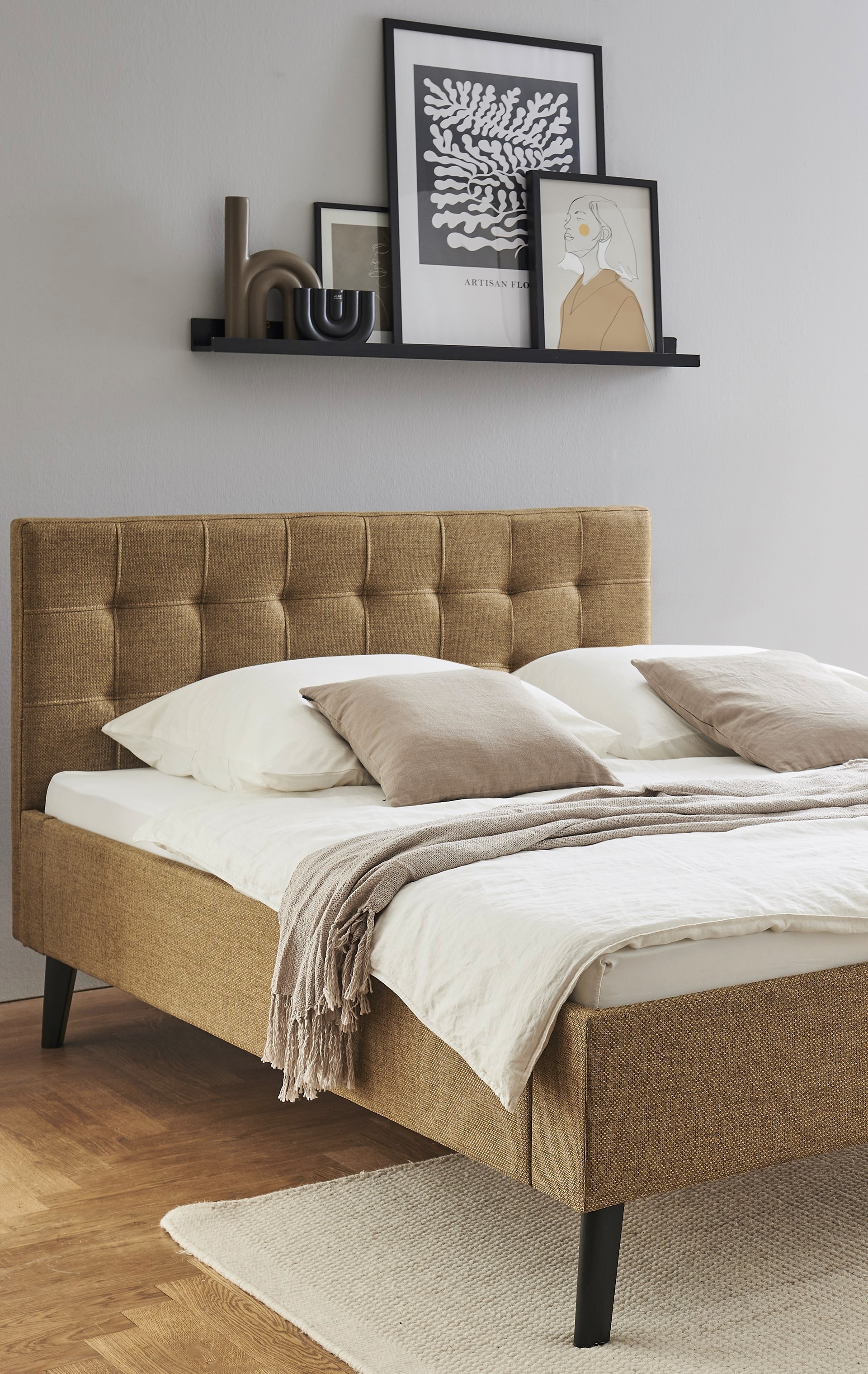 Modernes Schlafzimmer mit einem beigen Polsterbett, weißer Bettwäsche und grauen Kissen. Kunst und Dekoration befinden sich auf einem Regal über dem Bett, an einer grauen Wand.