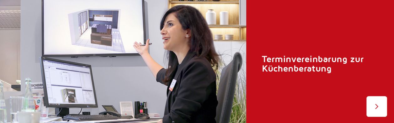 Eine Frau präsentiert an einem Schreibtisch einen Küchenentwurf auf einem an der Wand befestigten Bildschirm. Darunter lädt ein deutscher Text zur Buchung einer Küchenberatung ein.