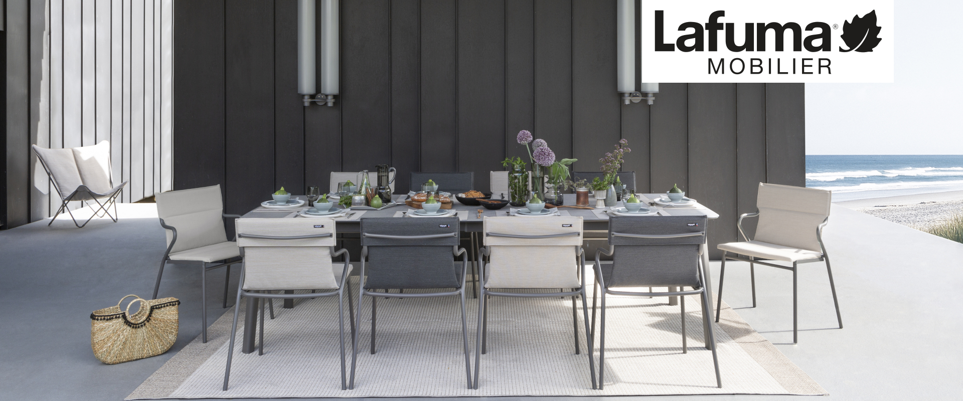 Eine Außenfläche am Strand mit dem großen Gartentisch Ancone von Lafuma Mobilier. Sein Alugestell und Platte in Zementoptik passen zu den Gartenstühlen der Serie in Sand beziehungsweise Schwarz.