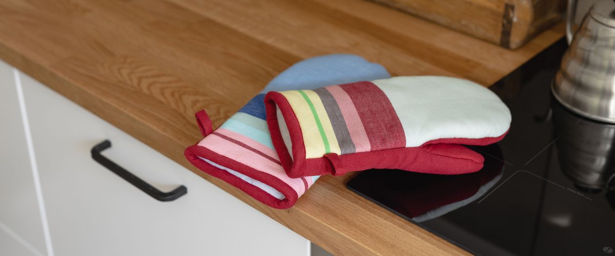 Die beiden Handschuhe des Ofenhandschuh Sets Nr. 1 von Remember Liegen auf eine hölzernen Arbeitsplatte. Sie haben ein schickes Design aus bunten Streifen.