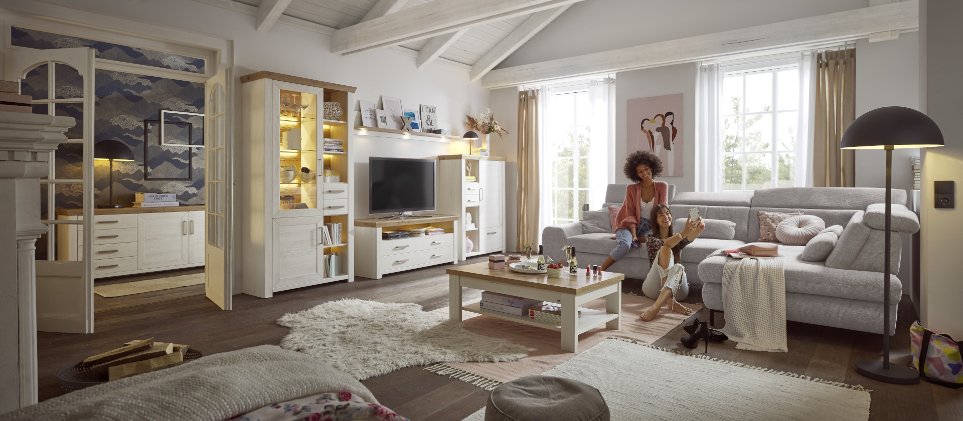 Die weiße Wohnzimmer Möbel Serie York im modernen Landhausstil mit Deckplatten im Look Eiche Artisan von set one by Musterring. Auf einem Sofa sitzen zwei junge Frauen und machen ein Selfie.