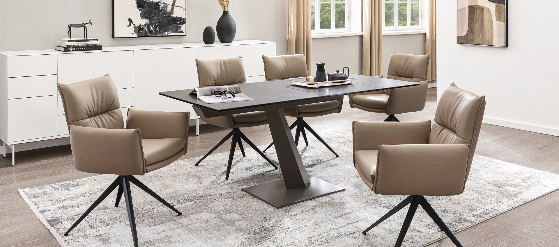 Modernes Esszimmer mit schwarzem, eckigem Esstisch und fünf beigen Lederstühlen. Ein hellgrauer Teppich und schlichte Deko runden das Raumkonzept ab.