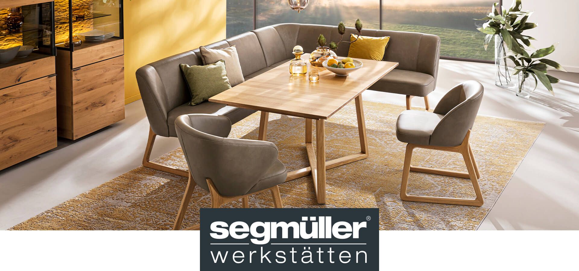 Die Speisegruppe Louis mit zwei Stuhlsesseln mit braunem Lederbezug und Holzgestell, einer dazu passenden Eckbank und einem Holztisch vor einer Lichtdurchfluteten Fensterfront